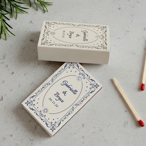 May include: Two white matchboxes with blue and cream decorative borders and text that reads "Gabrielle & Bryce" and a date. Two matches with red tips are also visible. The matchboxes are on a light gray surface.
