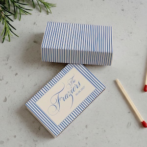 May include: Two rectangular matchboxes featuring a blue and white vertical striped pattern. One box displays the text The Fraziers and the date 06.06.2027 in blue script. Two wooden matches with red tips are placed next to the matchboxes.