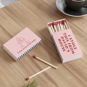 May include: Two pink matchboxes with red text and a wine glass graphic. One box says "COME BACK WITH WINE" and the other says "I STOLE THIS FROM DAVID'S HOUSE". Several matches with red tips are visible.