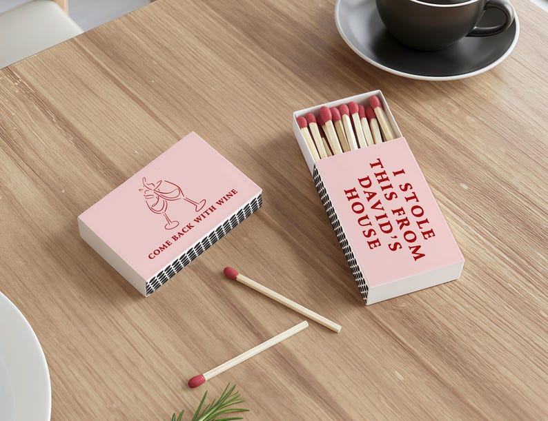 May include: Two pink matchboxes with red text and a wine glass graphic. One box says "COME BACK WITH WINE" and the other says "I STOLE THIS FROM DAVID'S HOUSE". Several matches with red tips are visible.