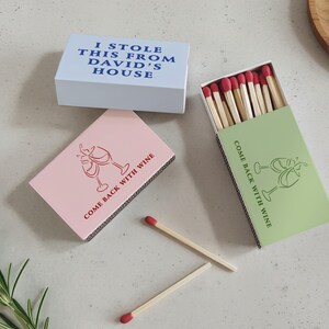 May include: Matchboxes in light blue, pink, and green. The light blue box reads "I STOLE THIS FROM DAVID'S HOUSE." The pink and green boxes feature a wine glass graphic and the text "COME BACK WITH WINE." The match heads are red.