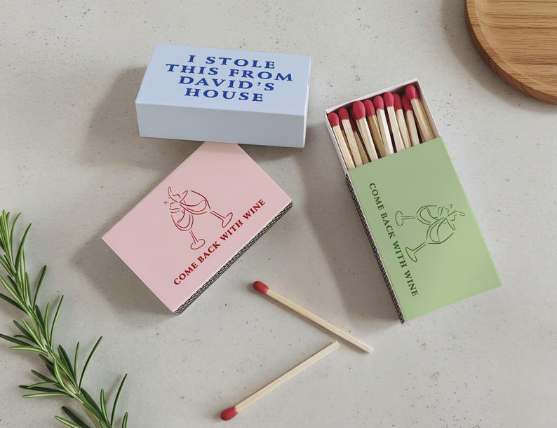 May include: Matchboxes in light blue, pink, and green. The light blue box reads "I STOLE THIS FROM DAVID'S HOUSE." The pink and green boxes feature a wine glass graphic and the text "COME BACK WITH WINE." The match heads are red.