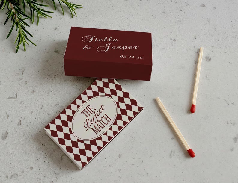 May include: A maroon matchbox with the names "Stella & Jasper" and the date "03.24.26" printed on it. A second matchbox with a white and maroon diamond pattern and the text "The Perfect Match". Two matches with red tips are also visible.