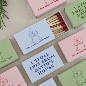 May include: Assortment of matchboxes in pastel colors, featuring wine glass illustrations and phrases like "Come Back With Wine" and "I Stole This From David's House." The matches have red tips.