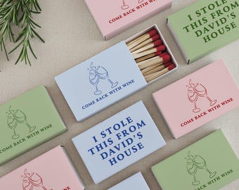 May include: Assortment of matchboxes in pastel colors, featuring wine glass illustrations and phrases like "Come Back With Wine" and "I Stole This From David's House." The matches have red tips.