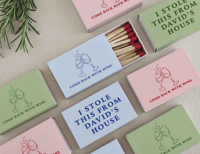 May include: Assortment of matchboxes in pastel colors, featuring wine glass illustrations and phrases like "Come Back With Wine" and "I Stole This From David's House." The matches have red tips.