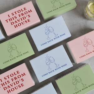 May include: A collection of small, rectangular boxes in various colors, including pink, green, and white. Some boxes feature the text "I STOLE THIS FROM DAVID'S HOUSE" in red. Other boxes have wine glass illustrations and the phrase "COME BACK WITH WINE."