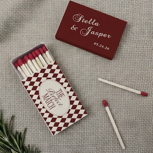 May include: A burgundy matchbox and a matching lid with the names "Stella & Jasper" and a date. The matchbox has a red and white diamond pattern and the text "The Perfect Match". Several matches are visible.