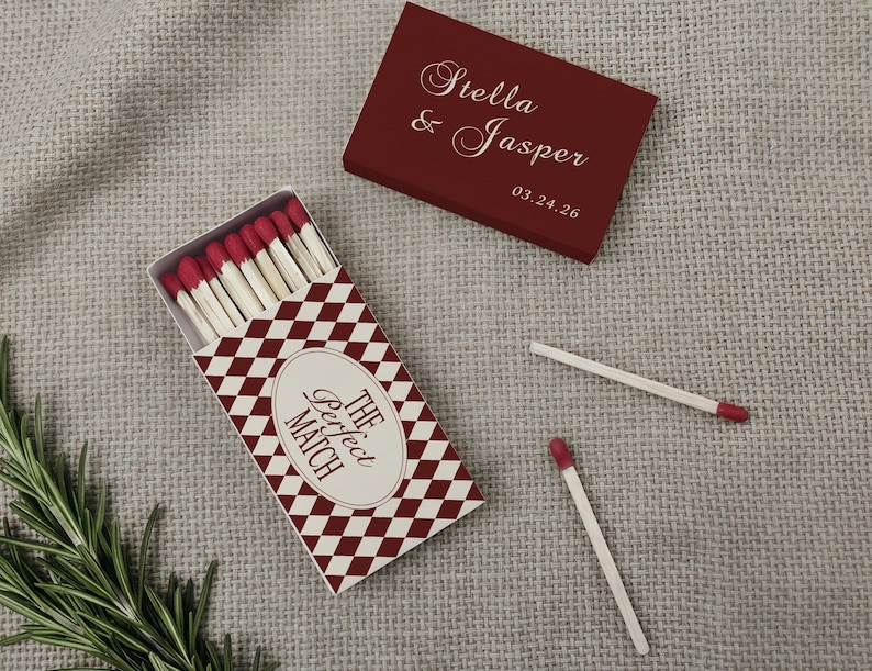 May include: A burgundy matchbox and a matching lid with the names "Stella & Jasper" and a date. The matchbox has a red and white diamond pattern and the text "The Perfect Match". Several matches are visible.