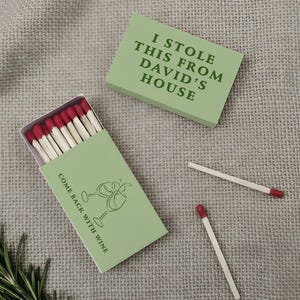 May include: A light green matchbox with the text "I STOLE THIS FROM DAVID'S HOUSE" and another with "COME BACK WITH WINE" and a wine glass graphic. The matches have red tips. A sprig of rosemary is in the corner.