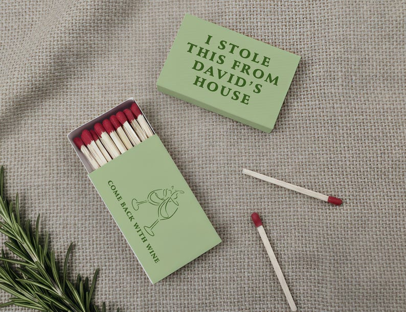 May include: A light green matchbox with the text "I STOLE THIS FROM DAVID'S HOUSE" and another with "COME BACK WITH WINE" and a wine glass graphic. The matches have red tips. A sprig of rosemary is in the corner.