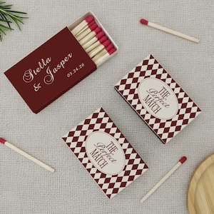 May include: Burgundy matchboxes with the names "Stella & Jasper" and the date "03.24.26" printed on them. Two additional matchboxes feature a diamond pattern and the text "The Perfect Match". Several matches with red tips are scattered around.