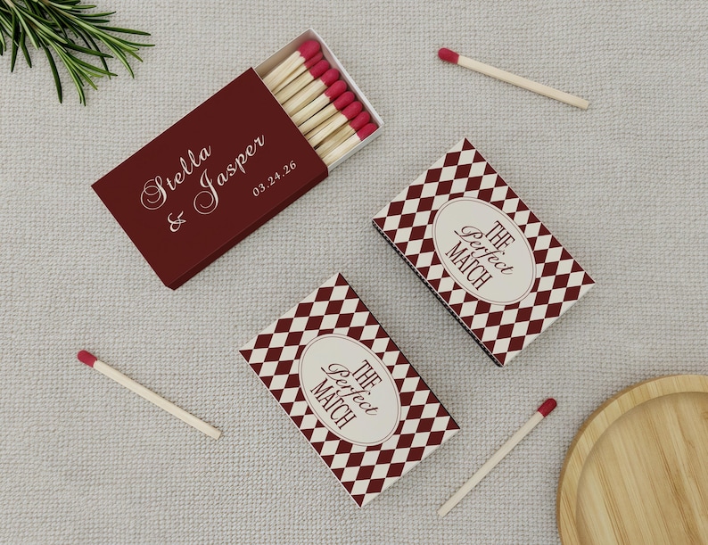 May include: Burgundy matchboxes with the names "Stella & Jasper" and the date "03.24.26" printed on them. Two additional matchboxes feature a diamond pattern and the text "The Perfect Match". Several matches with red tips are scattered around.