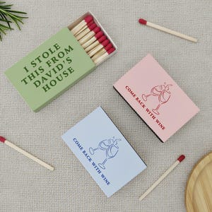May include: Matchboxes with red-tipped matches. One green box reads "I STOLE THIS FROM DAVID'S HOUSE." Two other boxes are pink and blue, with the text "COME BACK WITH WINE" and wine glass illustrations. A wooden tray and sprig of rosemary are also present.