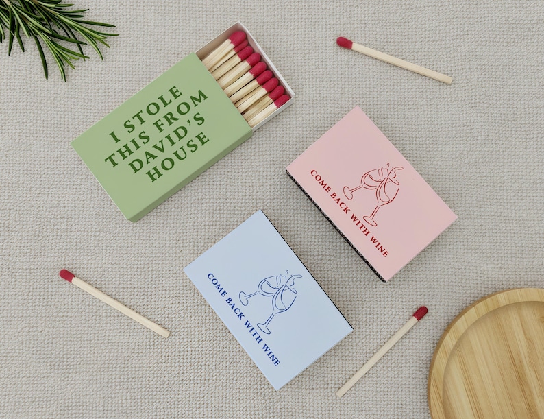 May include: Matchboxes with red-tipped matches. One green box reads "I STOLE THIS FROM DAVID'S HOUSE." Two other boxes are pink and blue, with the text "COME BACK WITH WINE" and wine glass illustrations. A wooden tray and sprig of rosemary are also present.
