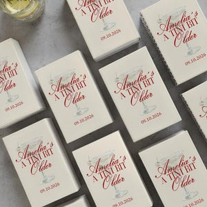May include: A collection of white gift boxes with the text "Amelia's A Tini Bit Older" in red script, along with the date 09.10.2026. A glass of water with lemon and rosemary is visible in the upper left corner.