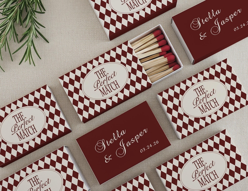 May include: Several matchboxes with a burgundy and white diamond pattern are arranged on a neutral surface. One box is open, revealing matches with red tips. The text "THE Perfect MATCH" is on the boxes. One box has the text "Stella & Jasper 03.24.26".