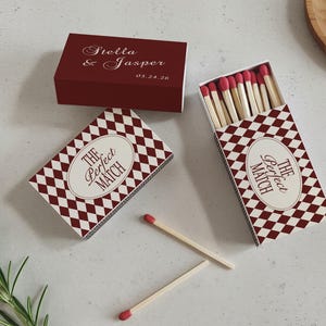 May include: Three matchboxes with a burgundy and white diamond pattern. One box is closed and has the names "Stella & Jasper" and a date. Two boxes are open, revealing matches with red tips. The text "The Perfect Match" is on the boxes.