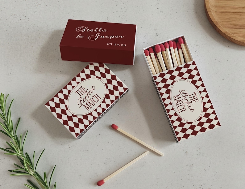 May include: Three matchboxes with a burgundy and white diamond pattern. One box is closed and has the names "Stella & Jasper" and a date. Two boxes are open, revealing matches with red tips. The text "The Perfect Match" is on the boxes.