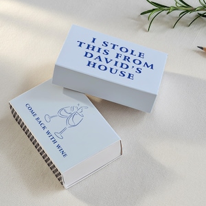 May include: Two white matchboxes with blue text and illustrations. One box reads "I STOLE THIS FROM DAVID'S HOUSE." The other reads "COME BACK WITH WINE" with wine glass illustrations. A pencil and rosemary sprig are in the background.