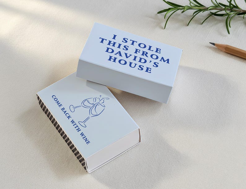 May include: Two white matchboxes with blue text and illustrations. One box reads "I STOLE THIS FROM DAVID'S HOUSE." The other reads "COME BACK WITH WINE" with wine glass illustrations. A pencil and rosemary sprig are in the background.