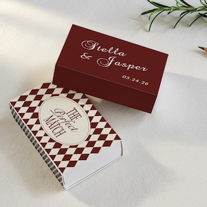 May include: Two decorative matchboxes. One is burgundy with the names "Stella & Jasper" and a date in white script. The other has a diamond pattern in burgundy and white, with the text "The Perfect Match" in an oval.