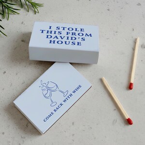 May include: Two white matchboxes with blue text and a wine glass design. One box reads "I STOLE THIS FROM DAVID'S HOUSE." The other says "COME BACK WITH WINE." Two matches with red tips are also visible.