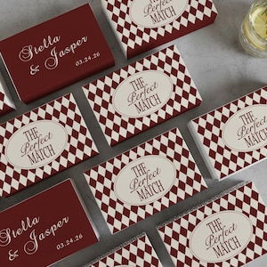 May include: A collection of matchboxes with a burgundy and cream diamond pattern. Some boxes feature the text "The Perfect Match" in an oval, while others display "Stella & Jasper" and a date. A refreshing drink is in the background.