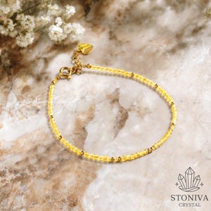 Könnte beinhalten: Ein zartes gelbes Perlenarmband mit goldfarbenen Akzenten. Das Armband hat kleine, runde gelbe Perlen und goldfarbene Abstandshalter. Der Verschluss und ein herzförmiger Anhänger sind ebenfalls goldfarben. Das Armband wird auf einer Marmoroberfläche präsentiert.