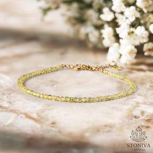 Puede incluir: Una delicada pulsera con pequeñas piedras preciosas de color amarillo claro ensartadas en una cadena y cierre dorados. La pulsera se exhibe sobre una superficie de mármol, con flores blancas en el fondo.