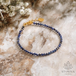 Może przedstawiać: Delikatna bransoletka z małymi, fasetowanymi niebieskimi koralikami nanizanymi razem. Bransoletka ma złote zapięcie i mały urok w kształcie serca. Tło to powierzchnia przypominająca marmur z białymi kwiatami.