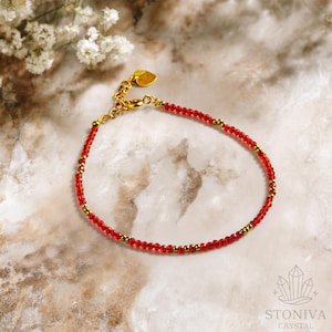 Puede incluir: Una delicada pulsera de cuentas rojas con detalles dorados y un dije en forma de corazón. La pulsera se exhibe sobre una superficie de mármol, con un logotipo en la esquina inferior derecha. La pulsera mide unos 18 cm de largo.