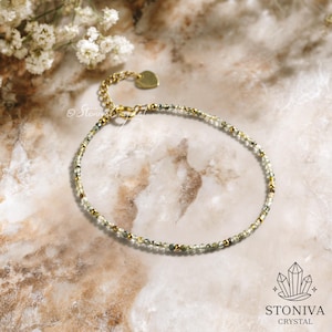 May include: A delicate gold bracelet featuring small, round, light green and gold beads. The bracelet has a heart-shaped charm and a clasp. The background is a marble-effect surface with white flowers.