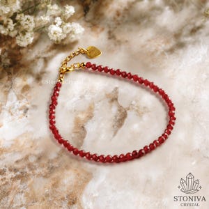 Puede incluir: Una delicada pulsera de cuentas rojas con una cadena dorada y un dije en forma de corazón. La pulsera está hecha de pequeñas cuentas rojas facetadas. La cadena dorada es ajustable. La pulsera se exhibe sobre una superficie de mármol.