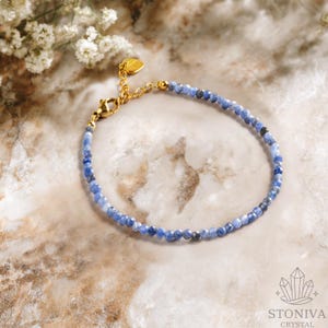Puede incluir: Una delicada pulsera con pequeñas piedras preciosas azules facetadas. La pulsera tiene un cierre dorado y un pequeño dije en forma de corazón. El fondo es una superficie clara y veteada con una ramita de flores blancas.