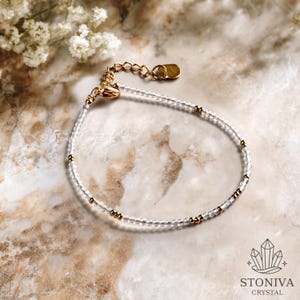 Puede incluir: Una delicada pulsera con cuentas de cristal transparente y detalles dorados. La cadena ajustable es dorada. La pulsera se muestra sobre una superficie de mármol. La marca "STONIVA CRYSTAL" es visible en la esquina inferior derecha.