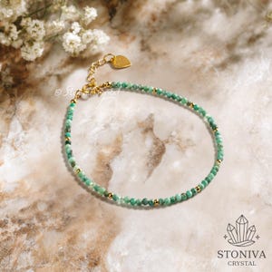 May include: A delicate emerald green beaded bracelet with gold accents. The bracelet features small, round green beads interspersed with gold-coloured beads and a gold heart-shaped charm. The bracelet is displayed on a marble surface.