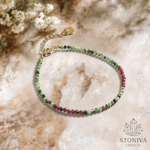 May include: A delicate bracelet featuring small, faceted beads in shades of green and burgundy. The bracelet has a gold-colored chain and clasp. The background is a marble-like surface with a floral accent.