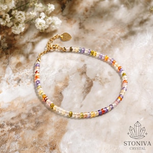 Könnte beinhalten: Ein zartes goldenes Armband, das mit einer Vielzahl farbiger Edelsteine verziert ist. Das Armband hat einen herzförmigen Anhänger und liegt auf einer Marmoroberfläche. Das Bild enthält auch den Text "STONIVA CRYSTAL".