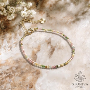 May include: A delicate bracelet with small, multi-coloured beads in pink, green, yellow, and brown. The bracelet has a gold-coloured chain and clasp. The background is a marble-like surface with white flowers.