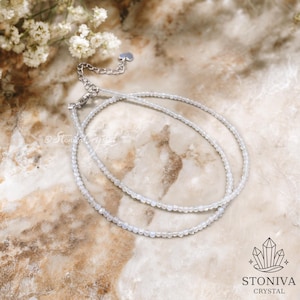 May include: A fine, double-strand necklace composed of small, round, pale beads. The necklace has a silver clasp and chain. The background is a marble-effect surface with white flowers. The brand name "STONIVA CRYSTAL" is visible in the lower right corner.