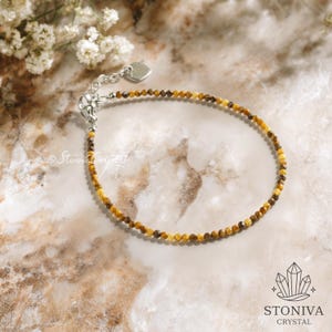 May include: A fine bracelet composed of small, amber-coloured beads. It has a silver-toned clasp and a tiny heart-shaped charm. The background is a marble-effect surface with a floral accent. The brand name "STONIVA CRYSTAL" is visible.
