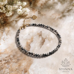 May include: A delicate bracelet featuring small, faceted black and gray beads strung together. The bracelet has a gold-colored chain and clasp, with a small heart-shaped charm. The background is a marble-like surface with white flowers.