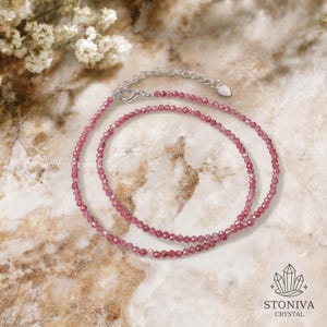 May include: A delicate pink gemstone necklace with a silver clasp. The necklace is composed of small, faceted beads, creating a sparkling effect. The background features a marble-like surface and floral accents. The brand name "STONIVA CRYSTAL" is visible.