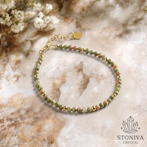 Puede incluir: Una pulsera de cuentas con piedras preciosas verdes y rojas y cuentas doradas. La pulsera es ajustable y tiene un cierre dorado. El fondo es una superficie de mármol con flores blancas.