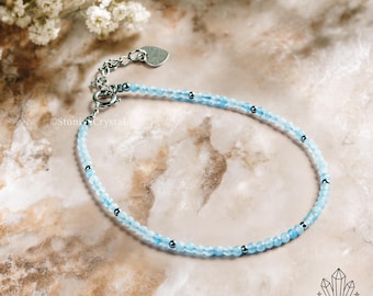 Puede incluir: Una delicada pulsera con pequeñas cuentas de color azul claro intercaladas con detalles plateados. La pulsera tiene una cadena plateada y un dije en forma de corazón. El fondo es una superficie similar al mármol con flores blancas.