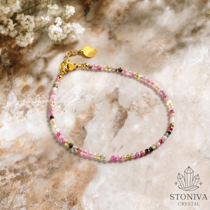 May include: A delicate bracelet with small, colourful gemstone beads in shades of pink, green, and black. The bracelet has a gold-coloured chain and clasp, with a small heart-shaped charm. The background is a marble surface with white flowers.