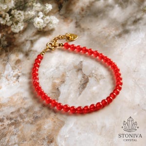 May include: A red beaded bracelet with a gold-coloured clasp and a small heart-shaped charm. The bracelet features small, round, red beads. The background is a marble-like surface with white flowers.