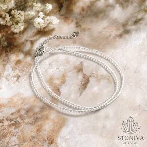 Op de afbeelding: Een delicate ketting van heldere kristallen kralen. De ketting is gemaakt van kleine, ronde, transparante kralen geregen op een zilveren ketting. De ketting wordt gepresenteerd op een marmeren oppervlak met witte bloemen op de achtergrond. Het Stoniva Crystal-logo staat rechtsonder.
