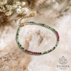 Könnte beinhalten: Ein zartes Armband mit kleinen, facettierten Perlen in Grün und Burgunderrot. Das Armband hat eine goldfarbene Kette und einen Verschluss. Der Hintergrund ist eine marmorähnliche Oberfläche mit floralen Akzenten.